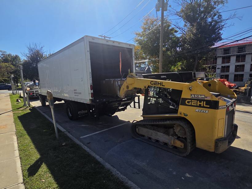 White box truck with its rear open on a New York street while a yellow Gehl RT215 skid steer loads a boxed pallet onto the lift gate