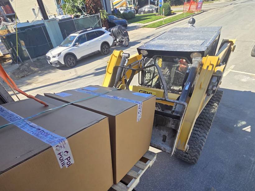 Skid steer operator moving two taped and strapped cartons on a pallet onto the back of the truck