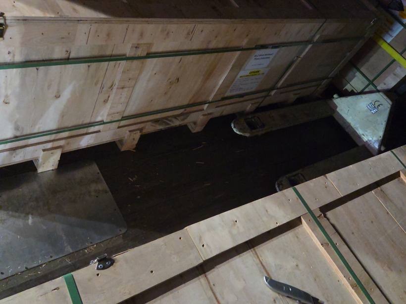 Long wooden freight crates strapped inside the dark cargo area of the box truck with a pallet jack nearby